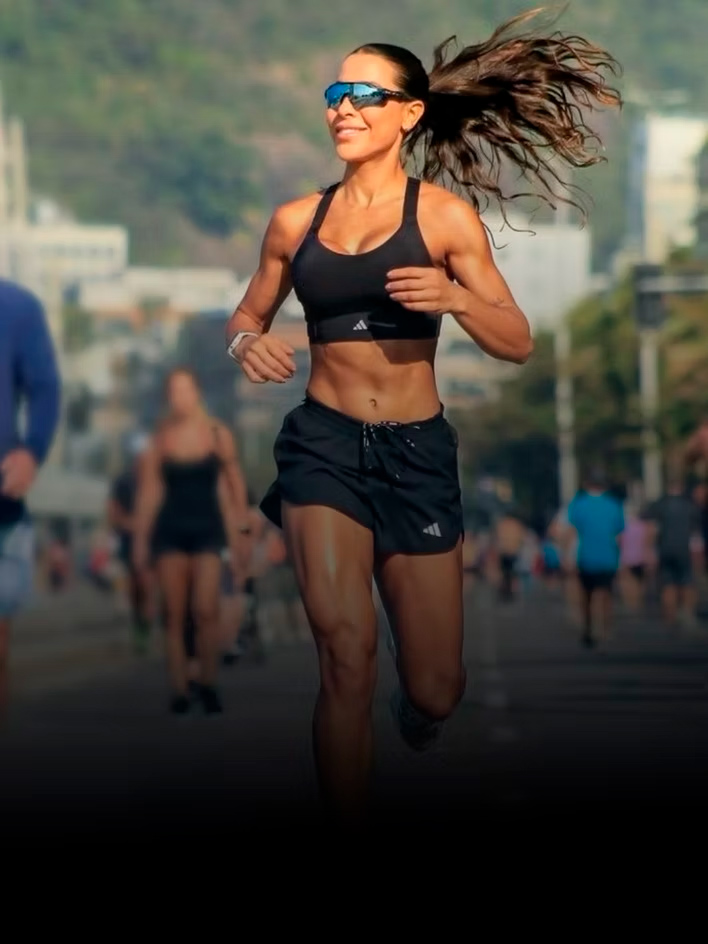 Mujer atlética sonriente corriendo al aire libre en una carrera urbana, usando un sujetador deportivo negro y gafas de sol, con el cabello al viento.